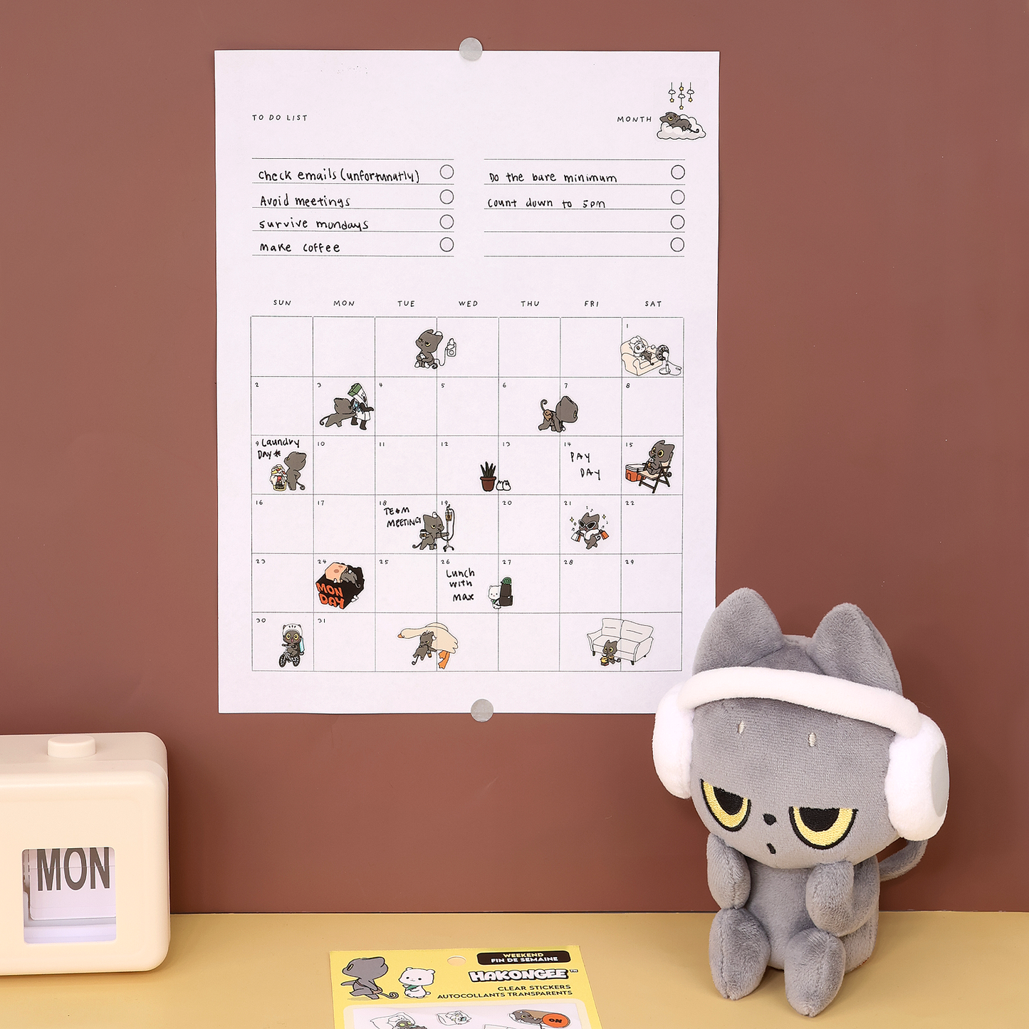 Hakongee wall calendar decorated with clear stickers beside a Hakongee grey cat plush and weekend sticker pack